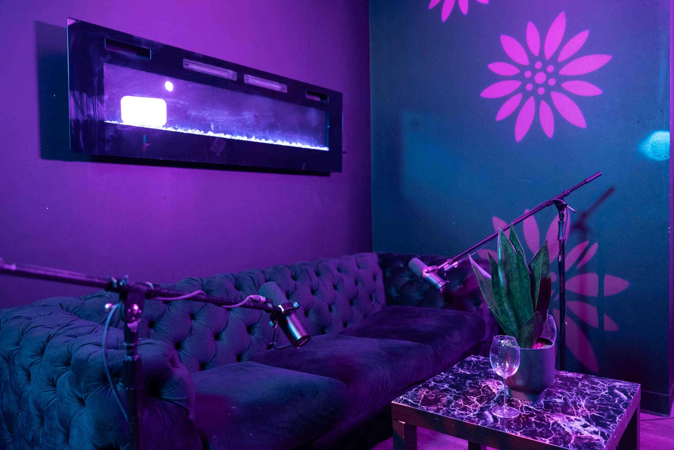 Angled view of the podcast lounge at Lush Event Space — fireplace, velvet sofa, boom mics, floral light projections
